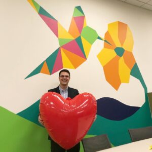 A photo of Brian Gialtketsis holding a large inflatable heart in front of a mural of a hummingbird